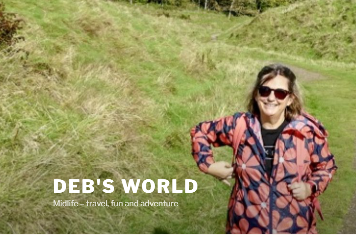 A confident midlife woman beams on a grassy trail, DEBS WORLD: Midlife – travel, fun & adventure for women over 40 shines below.